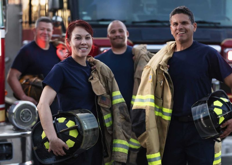 A multiracial group of four firefighters standing in front of two fire engines holding their helmets and fire protection suits, looking at the camera. The main focus is on the female firefighter, a Filipino woman in her 30s, in the foreground.