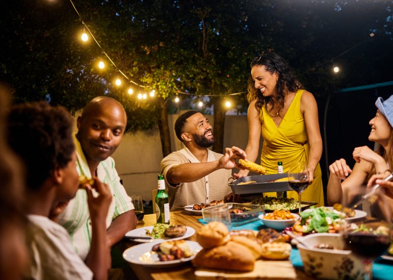 Family and friends dining at an outdoor table under string lights by the pool. Evening social gathering with food and drinks. Summer celebration concept. Medium shot.